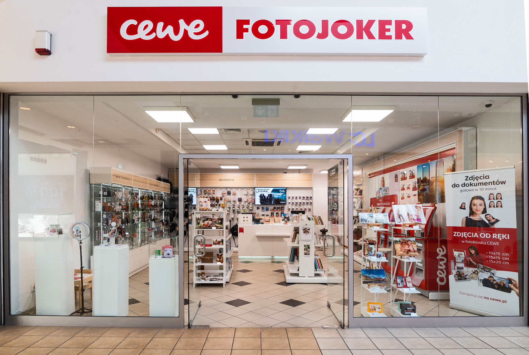 Wnętrze sklepu CEWE FOTOJOKER, w którym widoczne są wydruki fotograficzne, fotoksiążki, punkty samoobsługowe oraz gabloty z akcesoriami fotograficznymi.