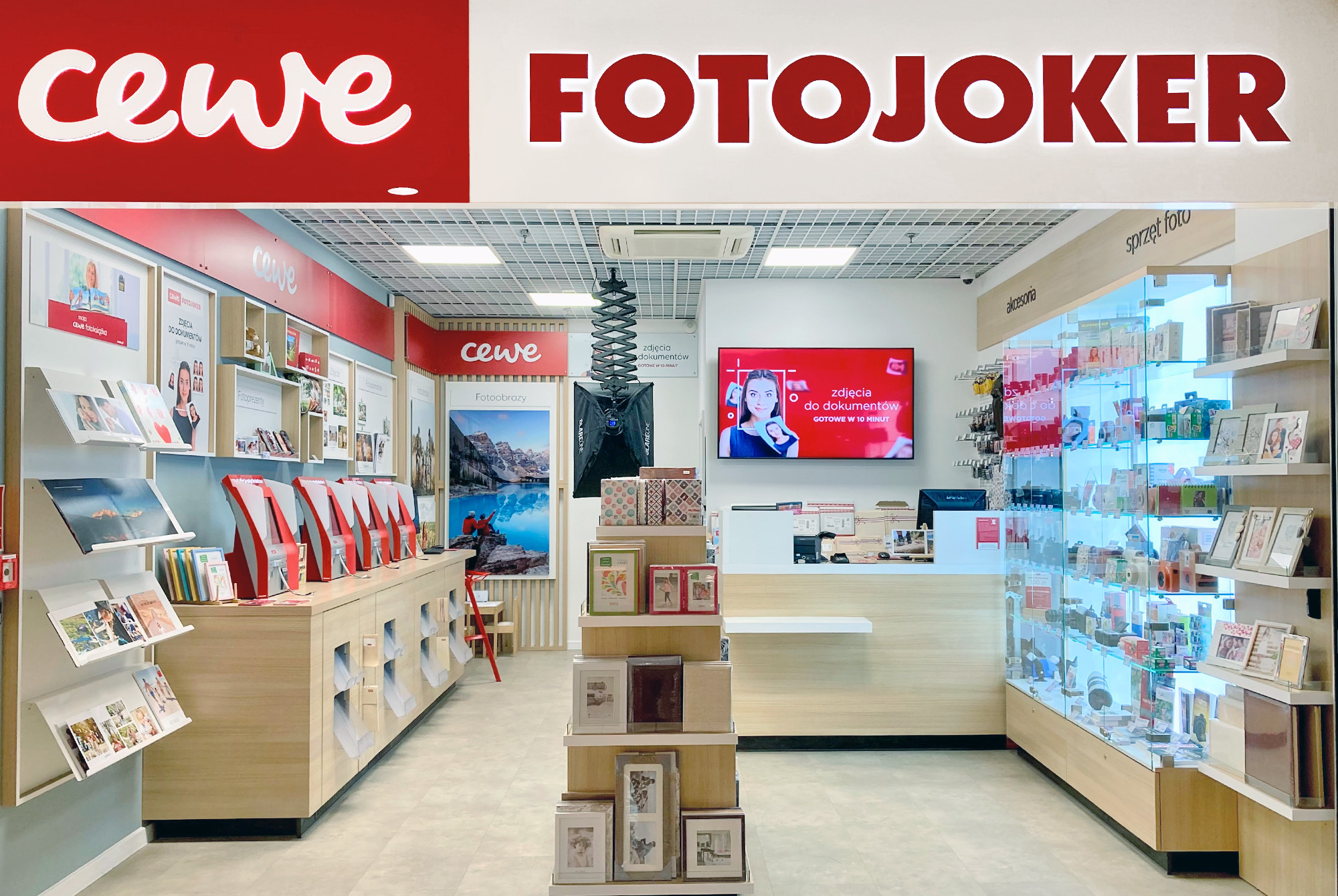 Wnętrze sklepu CEWE FOTOJOKER, w którym widoczne są wydruki fotograficzne, fotoksiążki, punkty samoobsługowe oraz gabloty z akcesoriami fotograficznymi.