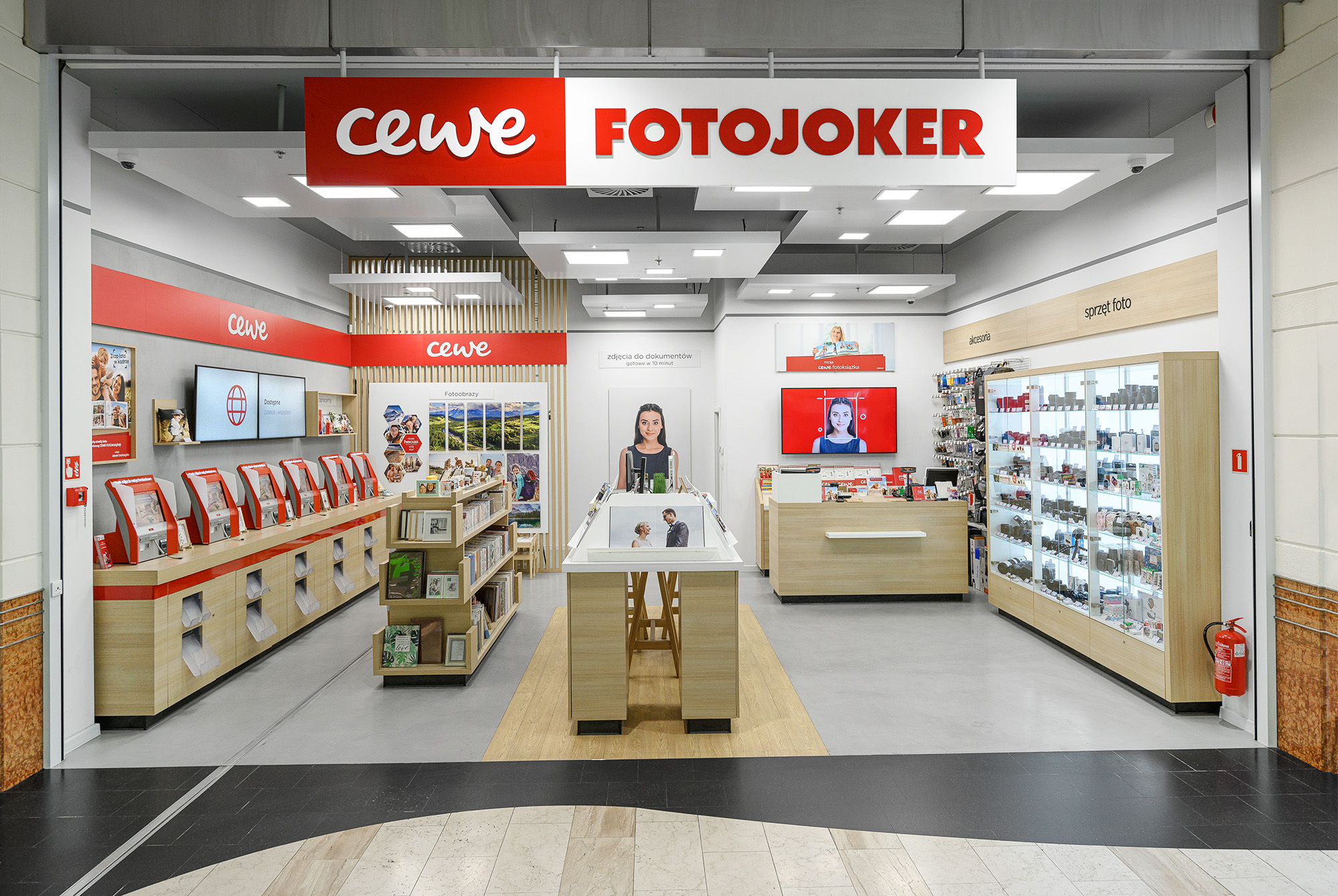 Wnętrze sklepu CEWE FOTOJOKER, w którym widoczne są wydruki fotograficzne, fotoksiążki, punkty samoobsługowe oraz gabloty z akcesoriami fotograficznymi.