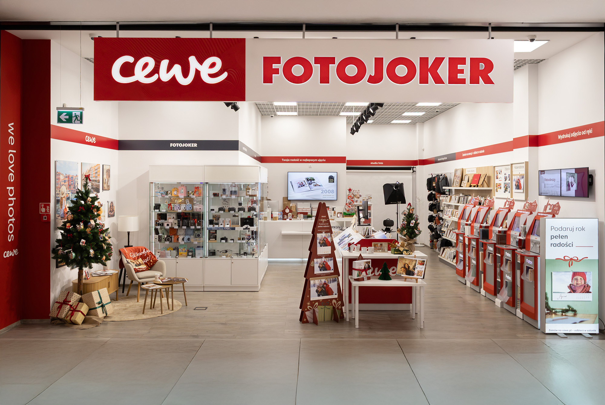 Wnętrze sklepu CEWE FOTOJOKER, w którym widoczne są wydruki fotograficzne, fotoksiążki, punkty samoobsługowe oraz gabloty z akcesoriami fotograficznymi.