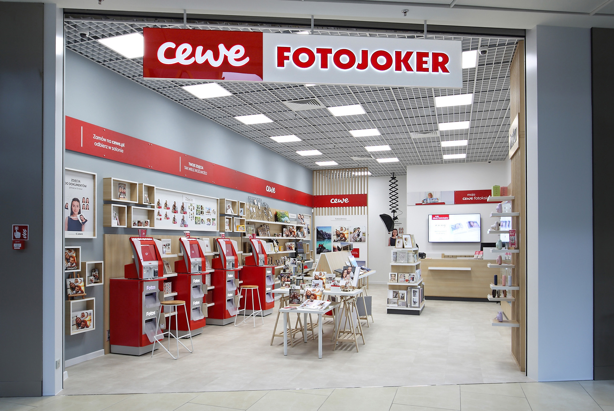 Wnętrze sklepu CEWE FOTOJOKER, w którym widoczne są wydruki fotograficzne, fotoksiążki, punkty samoobsługowe oraz gabloty z akcesoriami fotograficznymi.