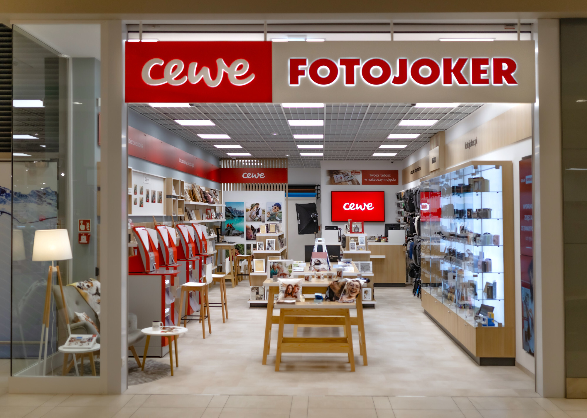Wnętrze sklepu CEWE FOTOJOKER, w którym widoczne są wydruki fotograficzne, fotoksiążki, punkty samoobsługowe oraz gabloty z akcesoriami fotograficznymi.