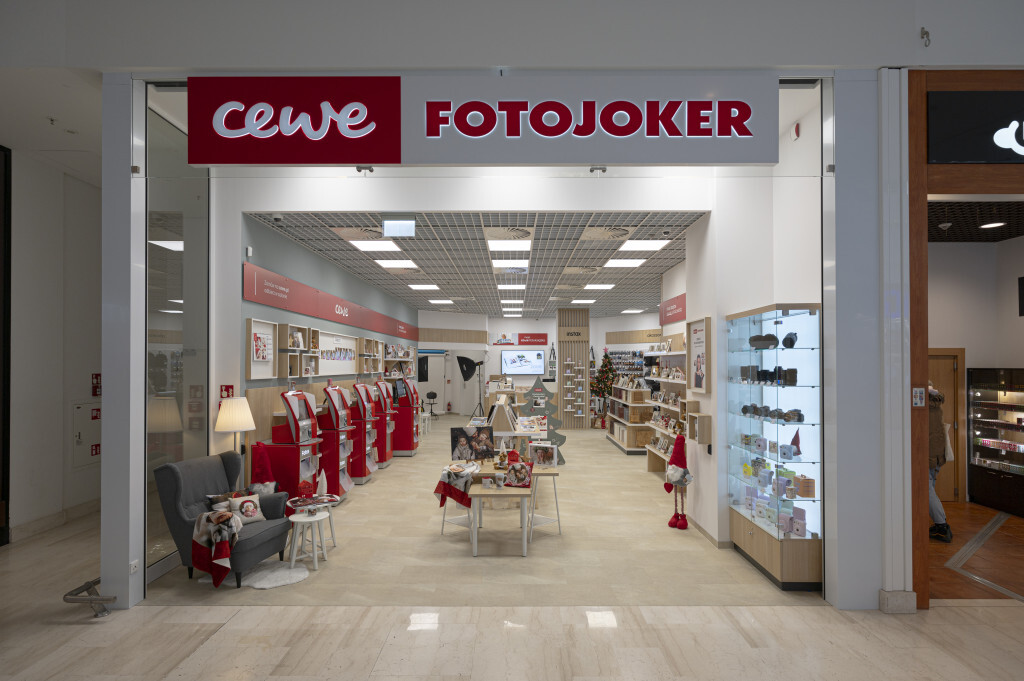 Wnętrze sklepu CEWE FOTOJOKER, w którym widoczne są wydruki fotograficzne, fotoksiążki, punkty samoobsługowe oraz gabloty z akcesoriami fotograficznymi.