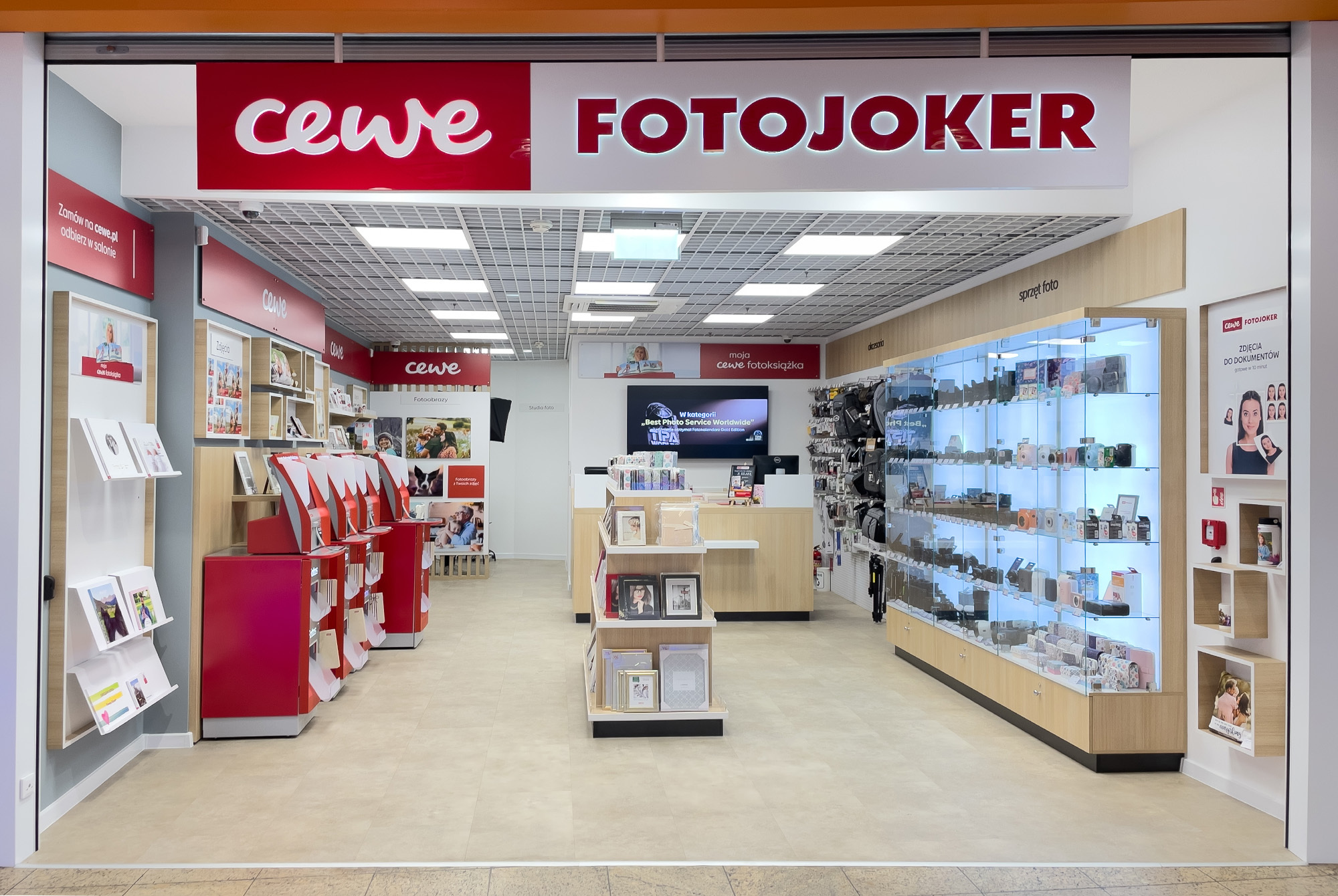 Wnętrze sklepu CEWE FOTOJOKER, w którym widoczne są wydruki fotograficzne, fotoksiążki, punkty samoobsługowe oraz gabloty z akcesoriami fotograficznymi.