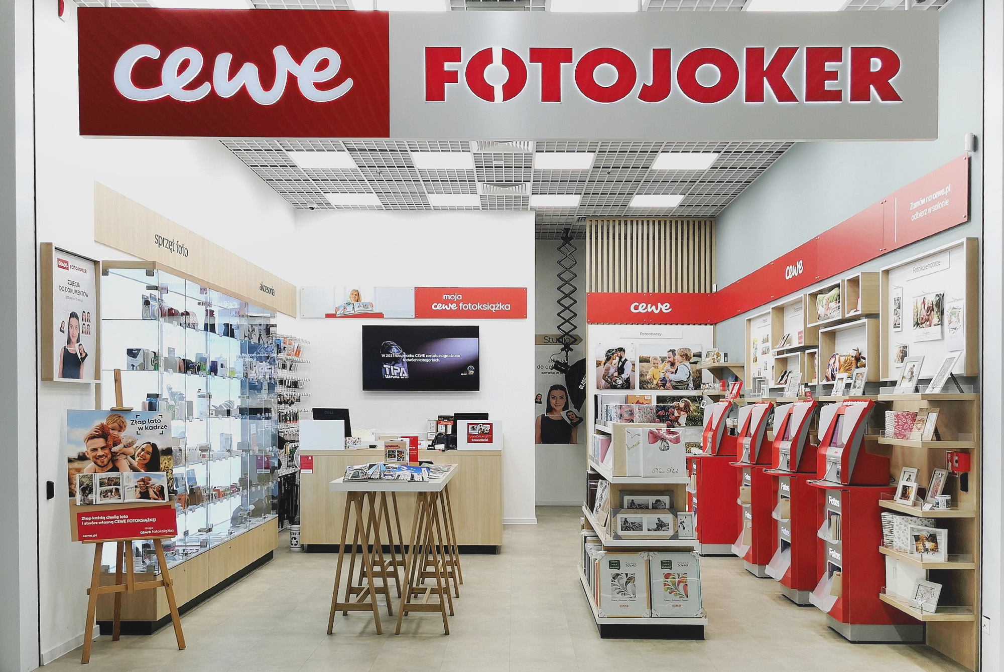 Wnętrze sklepu CEWE FOTOJOKER, w którym widoczne są wydruki fotograficzne, fotoksiążki, punkty samoobsługowe oraz gabloty z akcesoriami fotograficznymi.