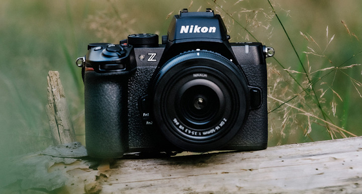 Aparat fotograficzny Nikon Z ustawiony na drewnianej powierzchni, w tle rozmyta roślinność Aparat fotograficzny Nikon Z ustawiony na drewnianej powierzchni, w tle rozmyta roślinność