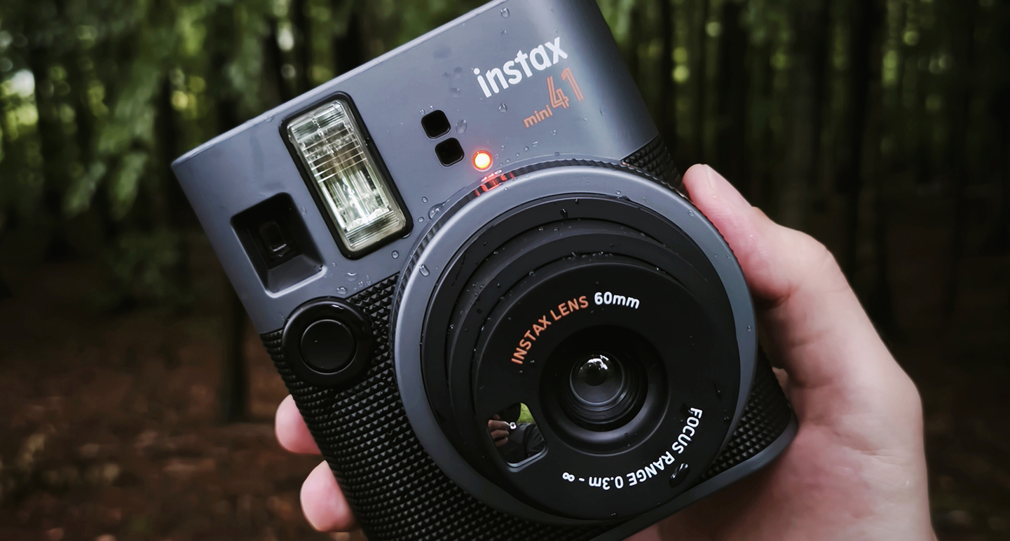 Aparat natychmiastowy Instax Mini 41, trzymany w dłoni w plenerze, prawdopodobnie w lesie. Obudowa aparatu jest ciemna, z widocznymi kroplami wody na powierzchni. Aparat natychmiastowy Instax Mini 41, trzymany w dłoni w plenerze, prawdopodobnie w lesie. Obudowa aparatu jest ciemna, z widocznymi kroplami wody na powierzchni.